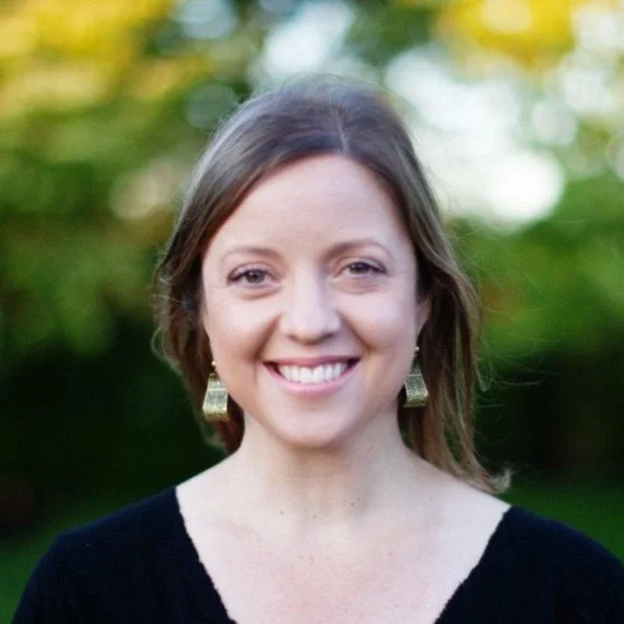 Smiling woman with light brown hair and earrings outdoors with green and yellow trees in the background.