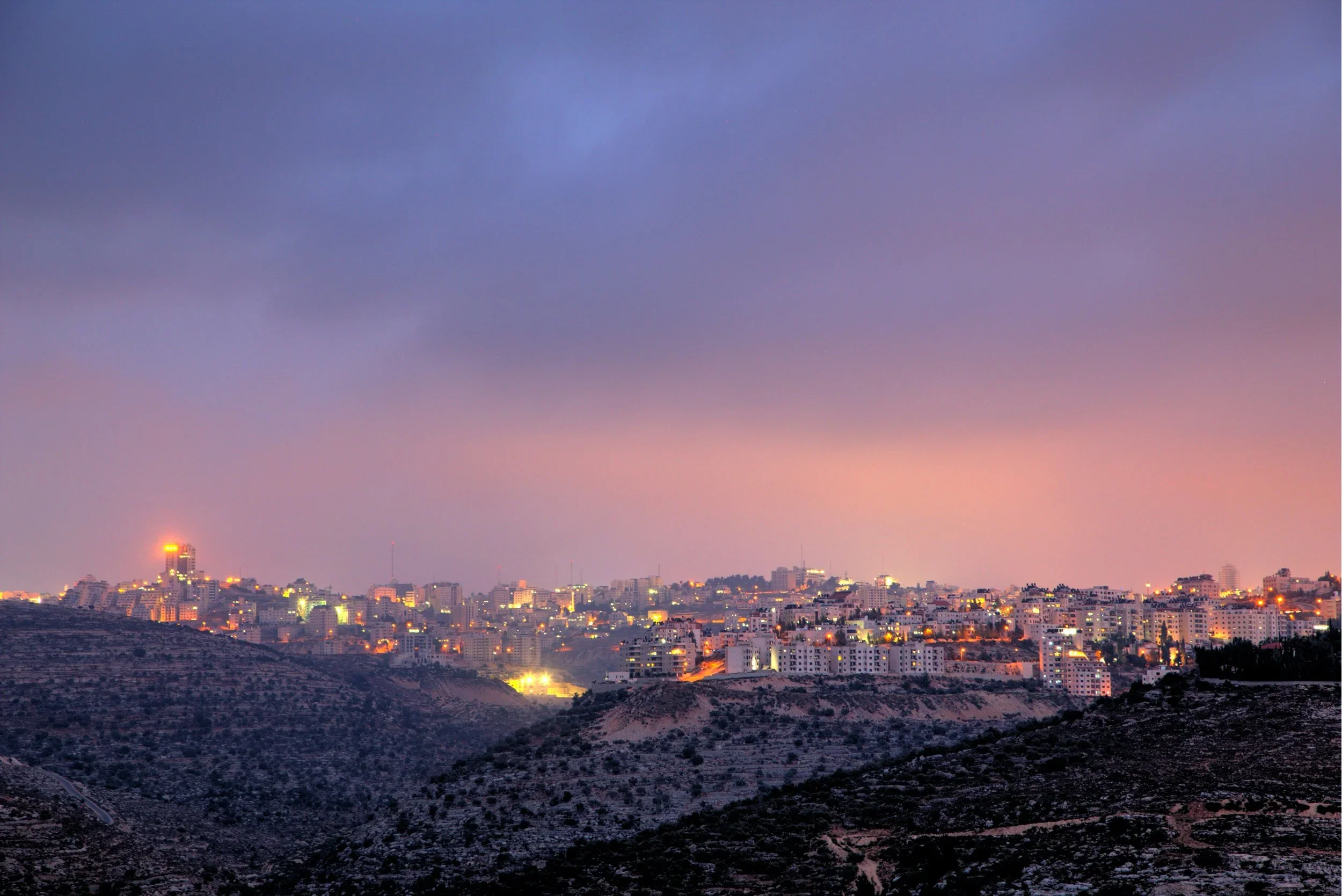 City skyline at dusk with illuminated buildings and a colorful sky with pink, purple, and blue hues.