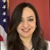 A woman with long dark hair smiling in front of a red background and an American flag.