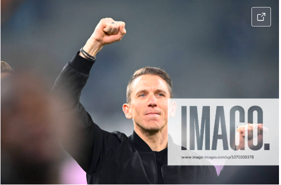 A man raising his fist in a gesture of victory or celebration, with a serious expression on his face, wearing a black jacket, in an outdoor setting.