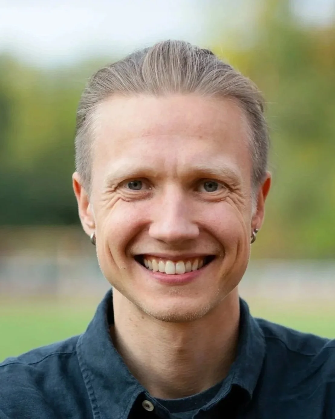 Close-up photo of a smiling person with short, slicked-back hair, wearing earrings and a dark-colored shirt, outdoors with blurred greenery in the background.