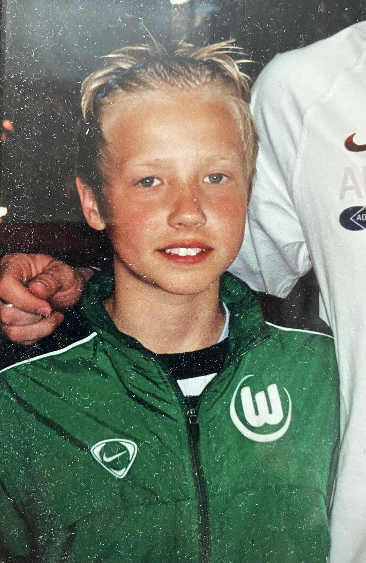 A young boy with blond hair and blue eyes smiling, wearing a green jacket with a white logo on the chest, standing next to a person in a white shirt.