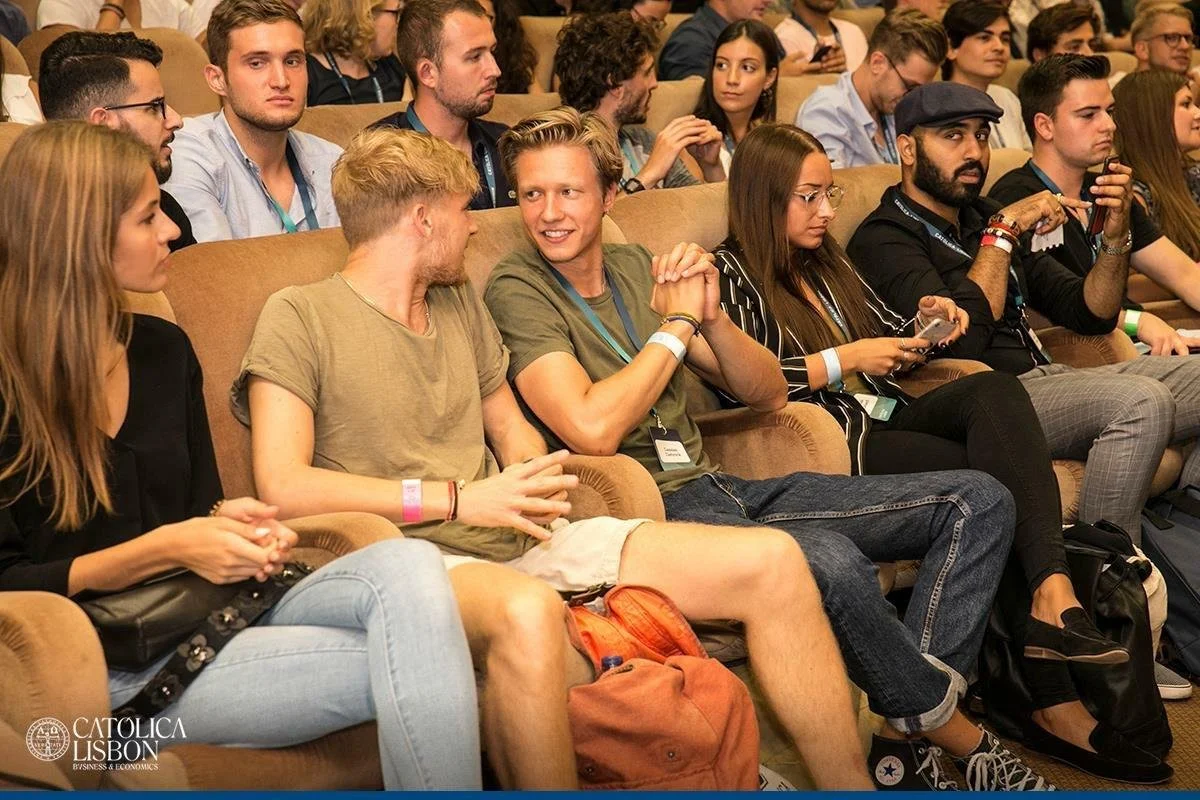 People seated in a conference room, some engaging in conversation and others looking at their phones, with a logo that reads 'CATOLICA LISBON BUSINESS & ECONOMICS' in the bottom left corner.