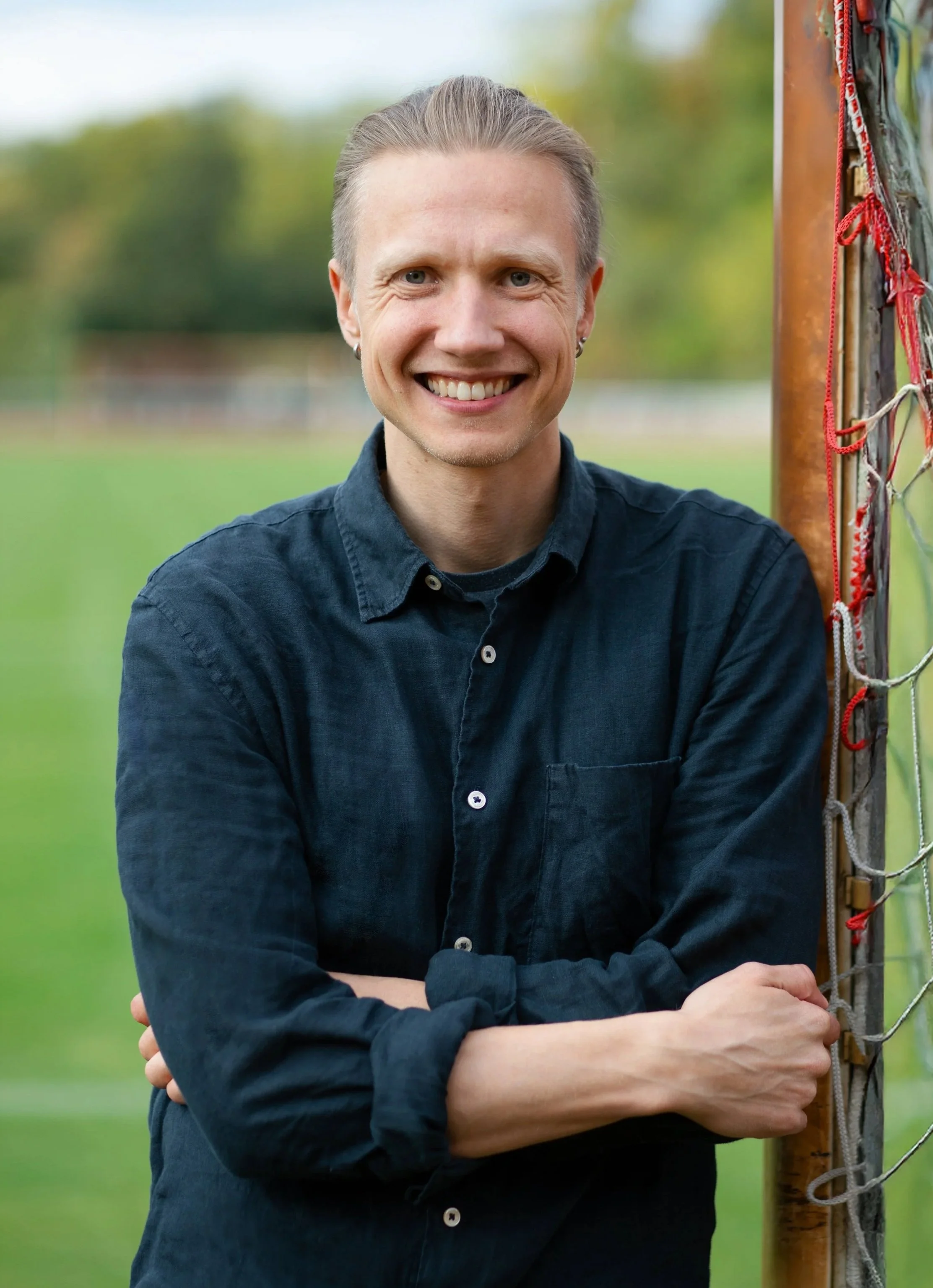 A smiling man with light skin, light brown hair, and blue eyes, wearing a dark blue button-up shirt, standing outdoors next to a soccer goalpost on a field with a blurred background of green grass and trees.