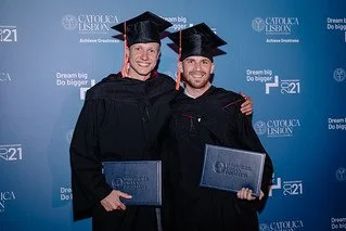 Two men in graduation caps and gowns holding diplomas, standing in front of a blue backdrop with logos and text.