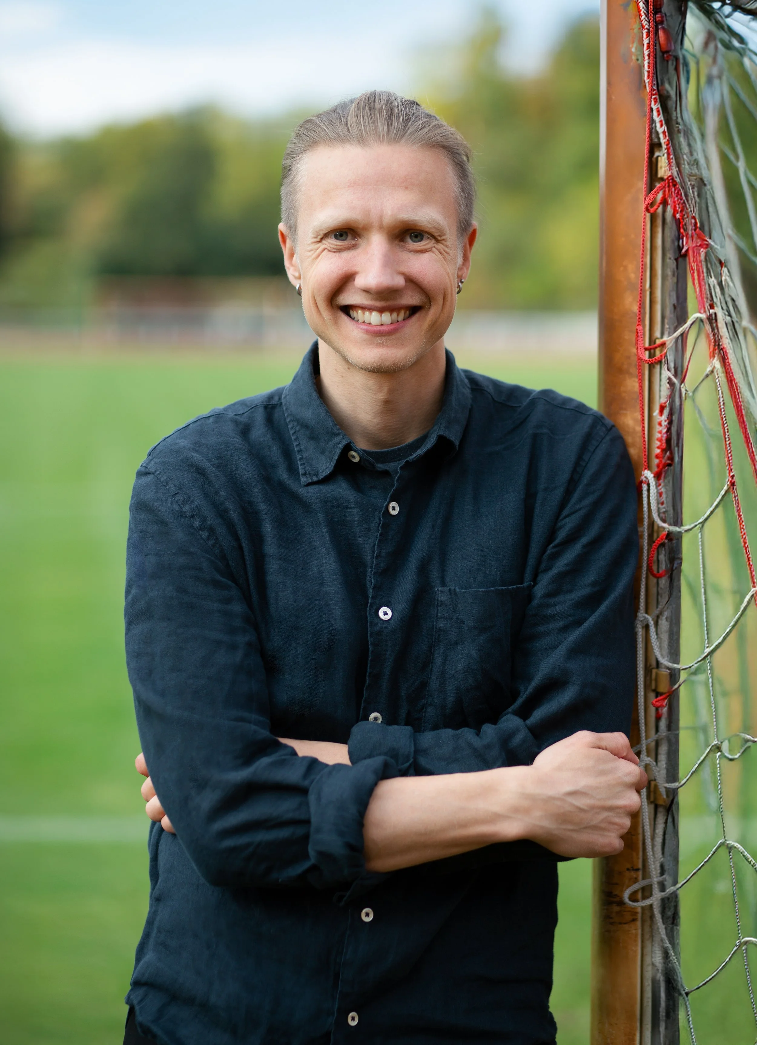 A smiling man stands outdoors next to a soccer goalpost on a grassy field. He is wearing a dark blue button-up shirt with sleeves rolled up and is clasping his arms across his chest.