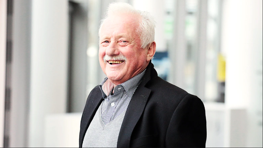 Smiling elderly man with white hair and mustache, wearing a black blazer over a gray sweater and checkered shirt, standing indoors with a blurred background.