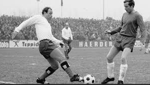 Two soccer players competing for the ball during a match, with one player in a white jersey and the other in a darker jersey, on a soccer field with spectators in the background.
