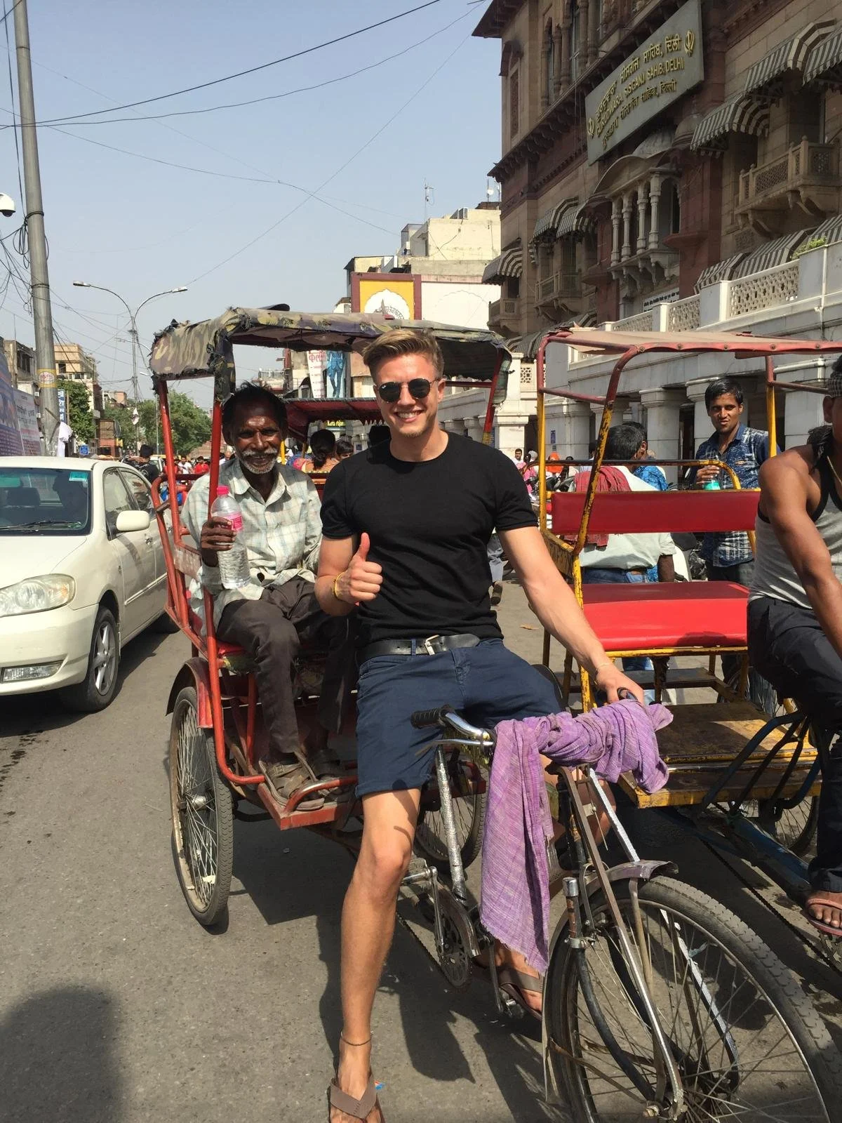 A young man with sunglasses and a black t-shirt, sitting on a cycle rickshaw with an older man in India, giving a thumbs-up. The busy street scene includes pedestrians and parked cars, with historic buildings in the background.
