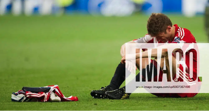A soccer player sitting on the field, holding his knee in pain with a uniform and cleats, red and white jersey, and a pile of gear nearby.