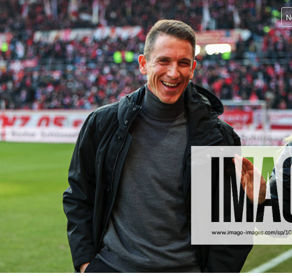 A smiling man wearing a black jacket over a gray turtleneck stands on a sports field with a large crowd in the background.