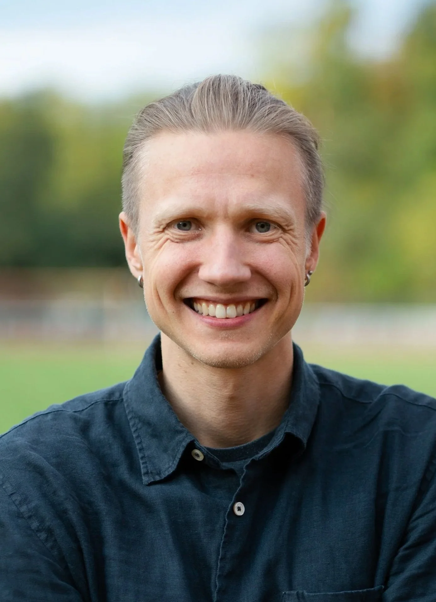 A smiling man with short, light brown hair, blue eyes, wearing earrings and a dark button-up shirt, outdoors with a blurred green and blue background.