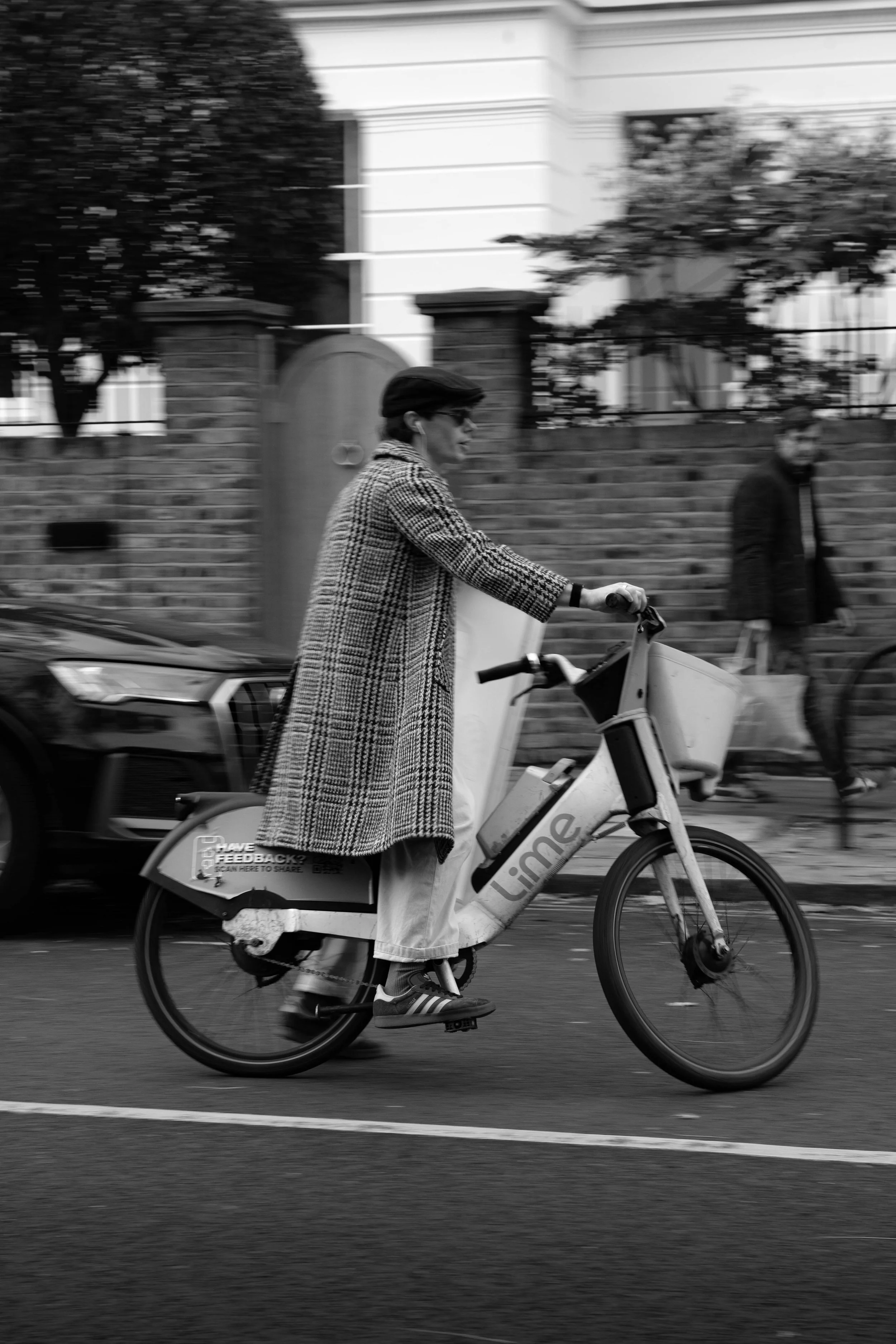 A woman rides a bike on a city street in black and white. She wears a checkered coat and glasses. A man walks in the background, carrying a shopping bag, alongside a brick wall and parked cars.