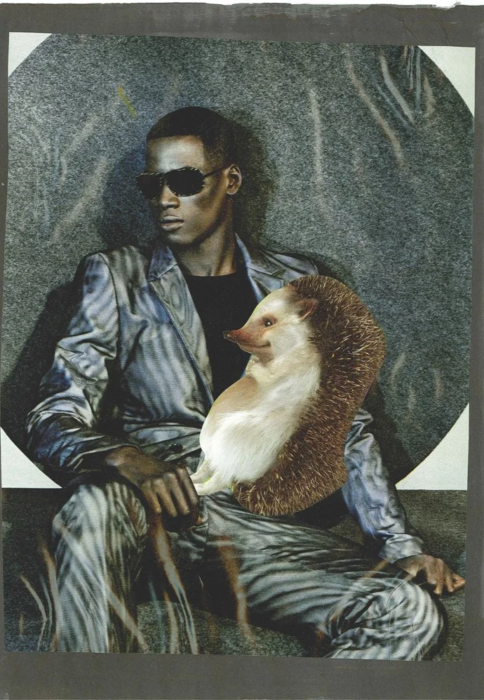 A woman wearing sunglasses and a shiny silver jacket, sitting with one hand on her leg, holding a hedgehog that has the body of a ferret with a hedgehog's face and quills, against a dark background.