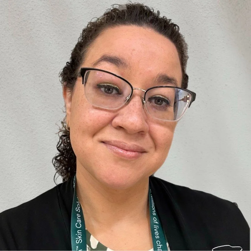 Close-up photo of a woman with dark curly hair, wearing glasses, a black shirt, and a teal lanyard, smiling at the camera against a plain background.