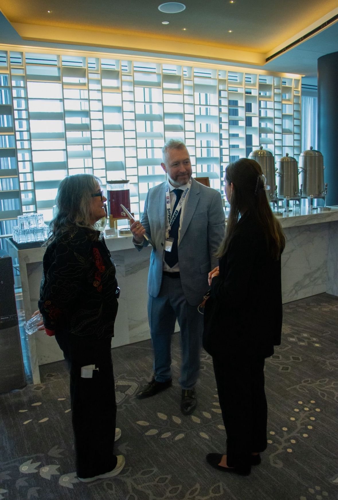 Three people in formal attire having a conversation at a conference or event in a modern, well-lit venue with a marble refreshment counter and coffee urns in the background.