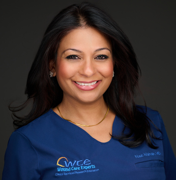 A smiling woman with long dark hair wearing a blue medical scrub top with a logo on it, against a dark background.