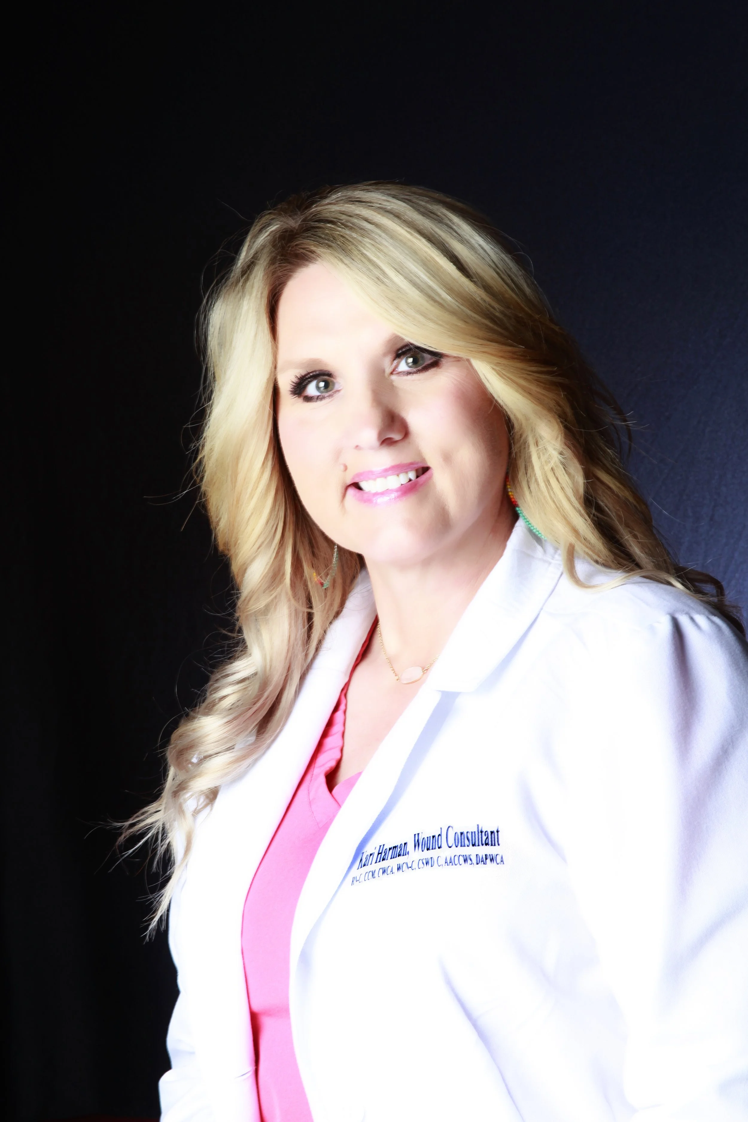 Professional portrait of a woman with blonde hair, wearing a white coat and pink top, against a dark background.