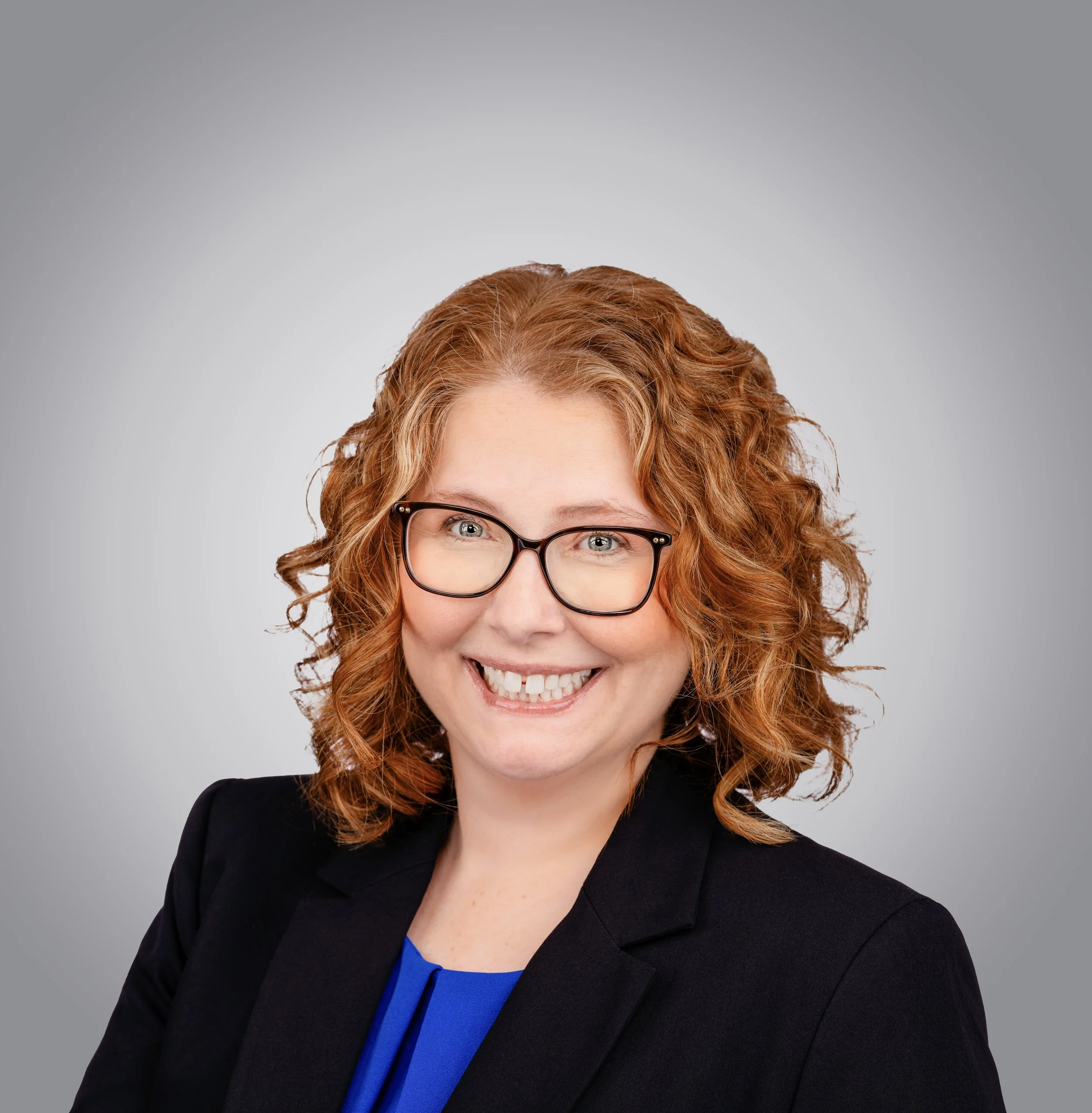 A woman with curly red hair, glasses, and a big smile, wearing a black blazer and a blue top against a gray background.