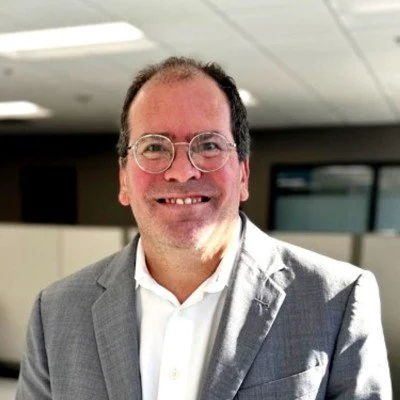 A man in a gray suit and white shirt smiling in an office setting.