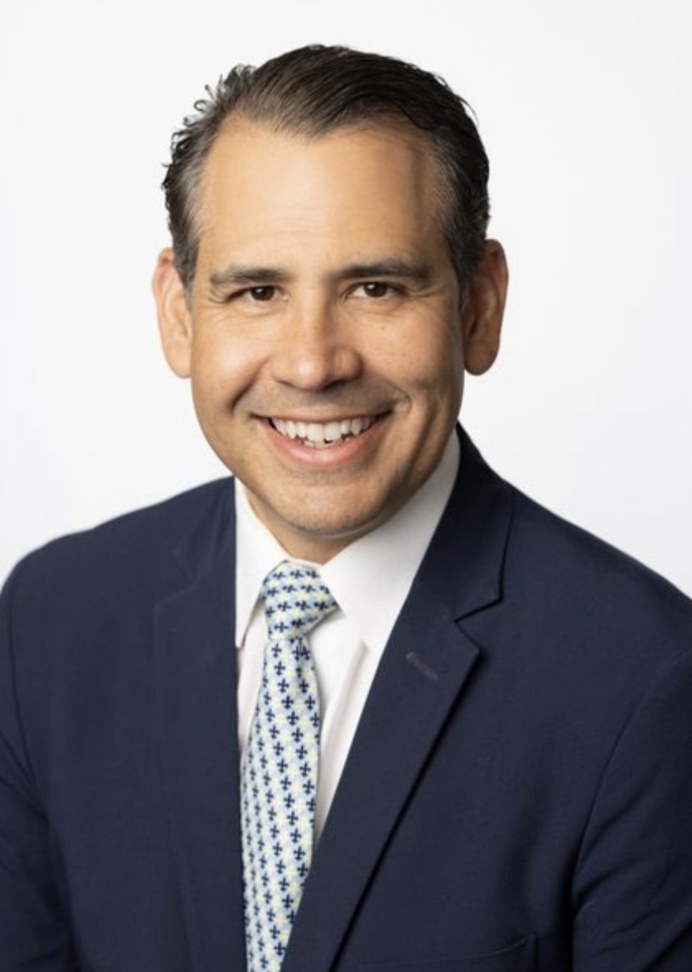 Professional headshot of a smiling man in a dark suit, white shirt, and patterned tie, with a plain white background.