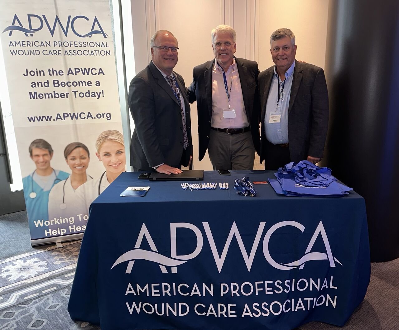 Three men standing behind a table with an ADWCA banner at an event or conference, with a sign promoting the American Professional Wound Care Association, featuring images of healthcare professionals.