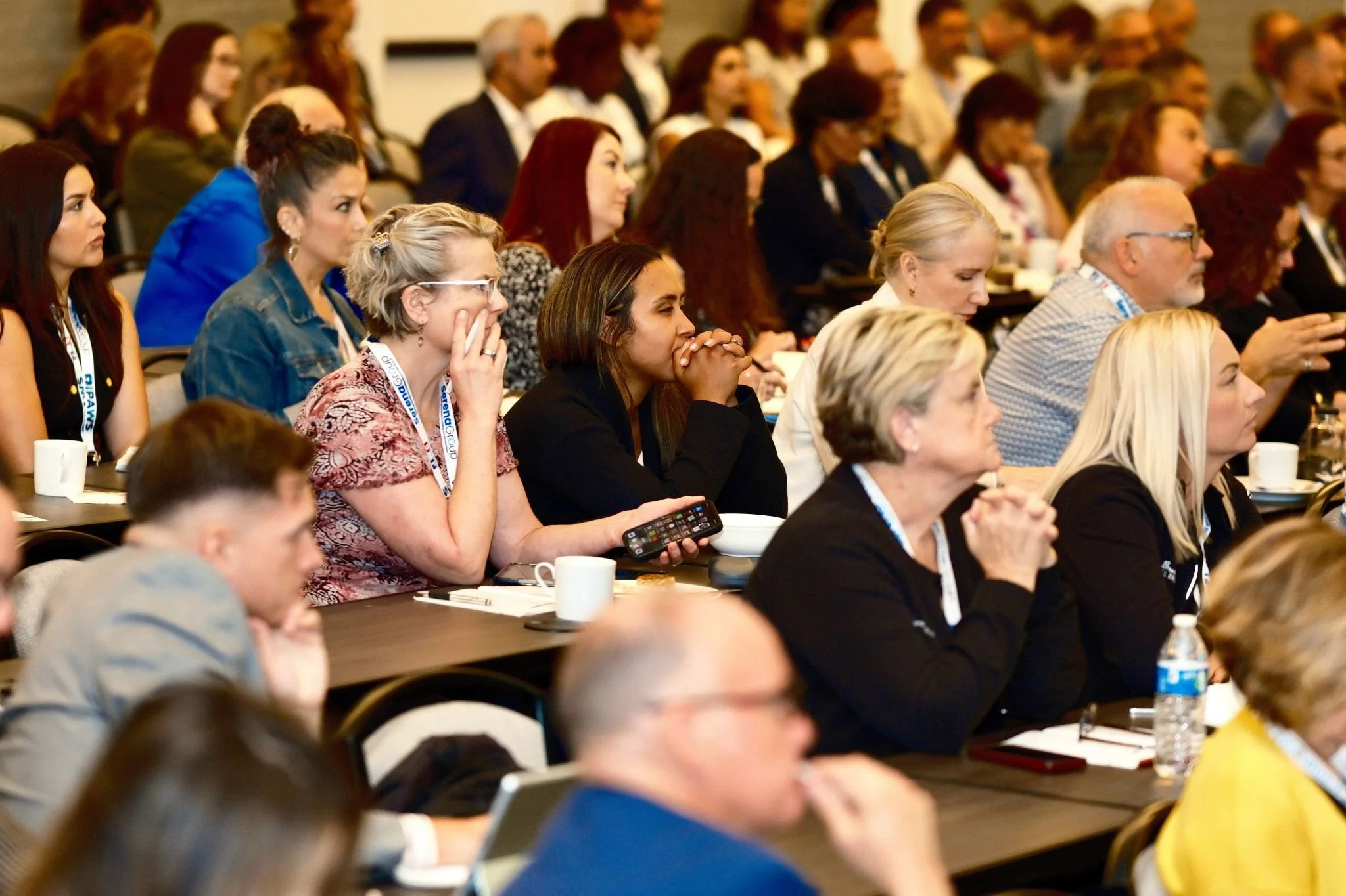 A large group of diverse people attending a conference or seminar, sitting at tables with notebooks, cups, and bottles, listening attentively.