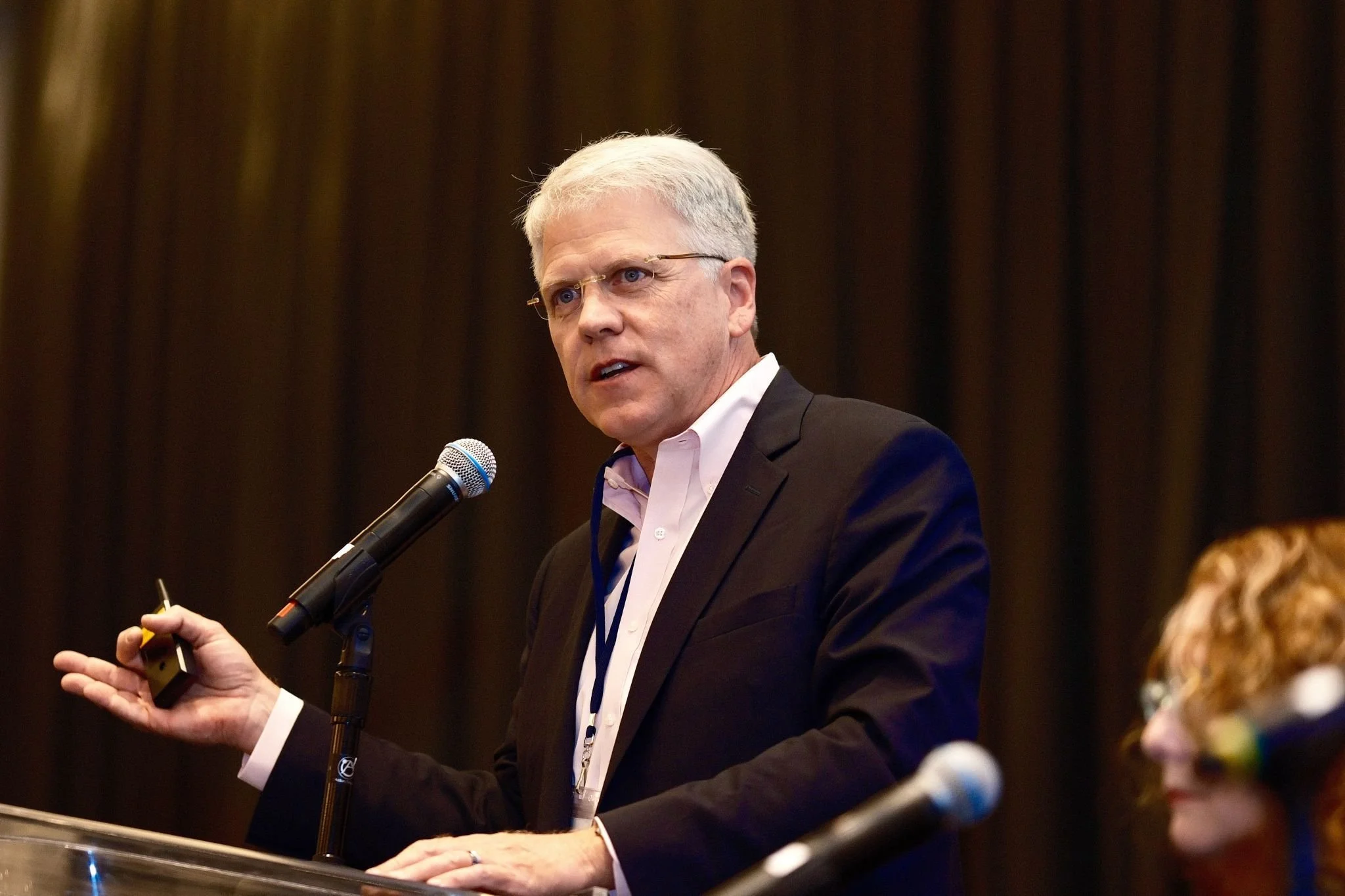 A man with gray hair and glasses giving a presentation at a conference, speaking into a microphone and holding a remote, with dark curtains in the background.