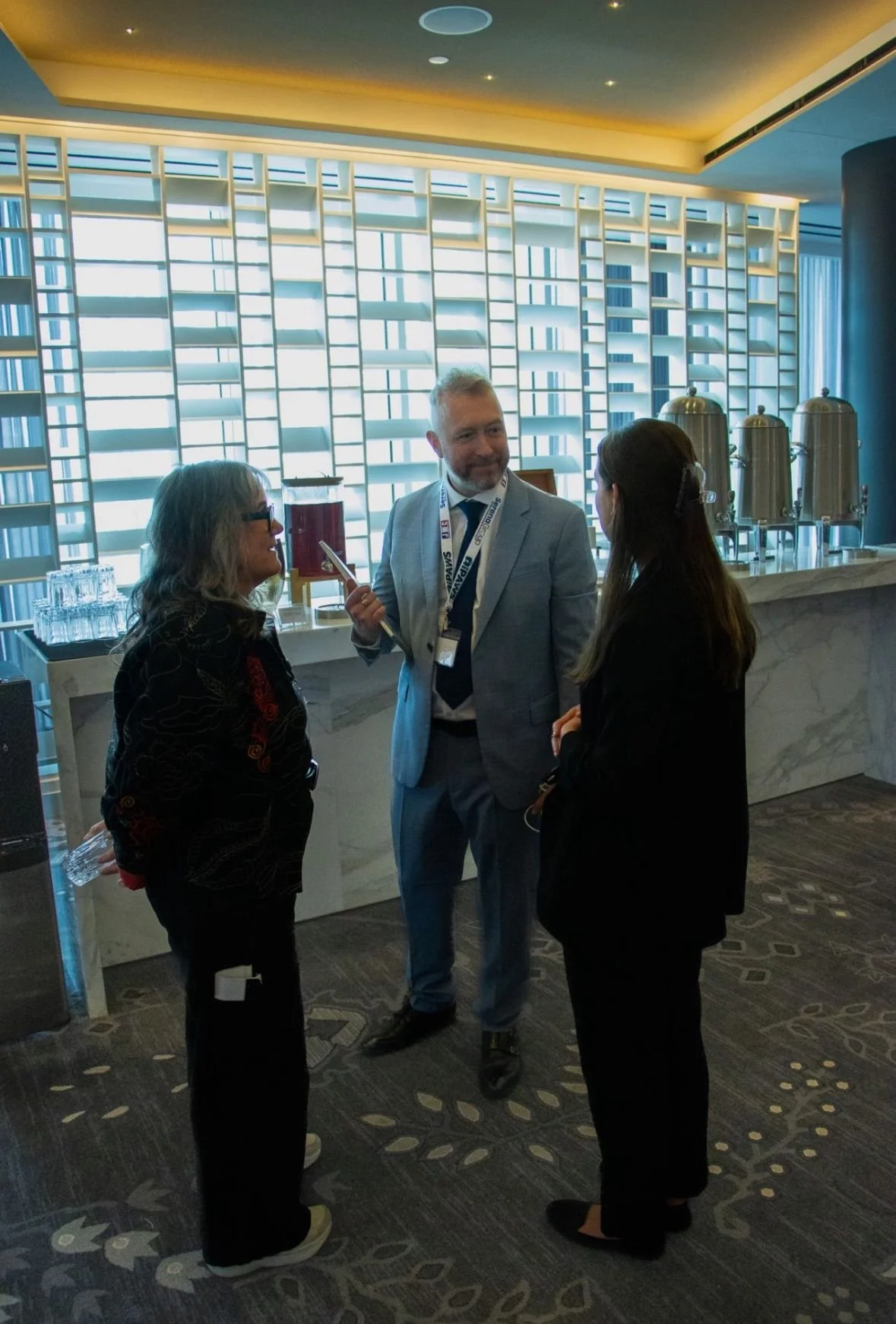 Three people having a conversation in a modern, well-lit indoor setting with a high ceiling, large windows with lattice-style blinds, and a marble counter with beverage dispensers in the background.