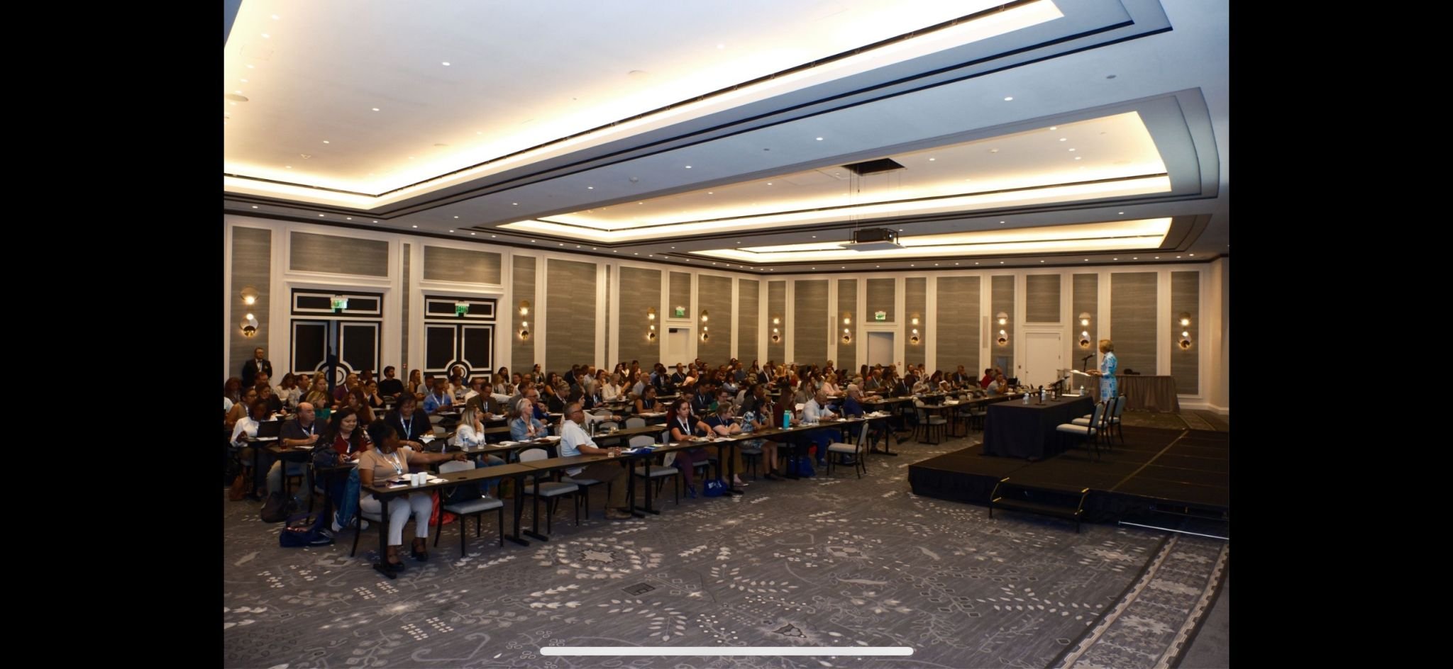 Conference room filled with attendees seated at tables, facing a stage where a speaker is presenting. Modern decor with wall sconces and high ceilings.