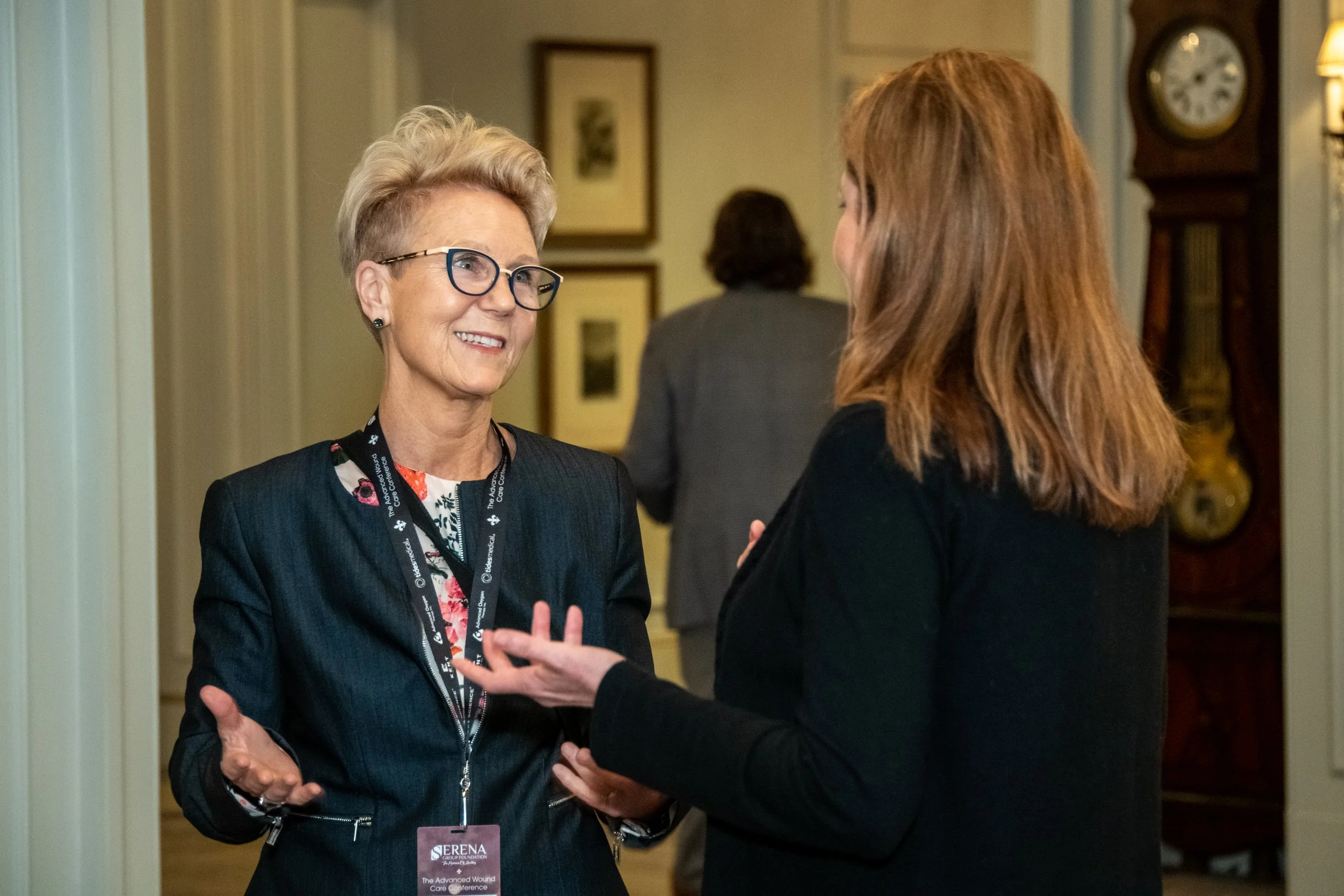 Two women talking at a formal event, one with short blonde hair and glasses, the other with long red hair, both smiling.