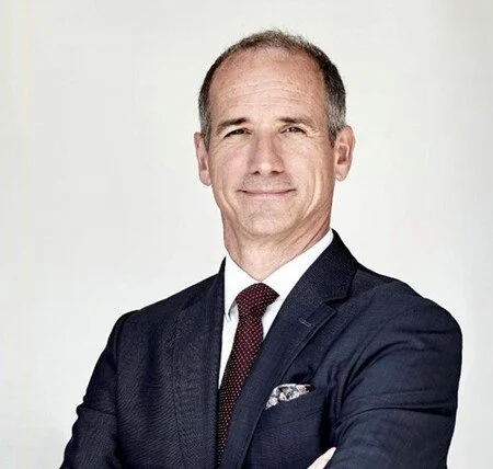 A middle-aged man with short hair, wearing a dark suit, white shirt, and red tie, smiling with arms crossed against a plain white background.