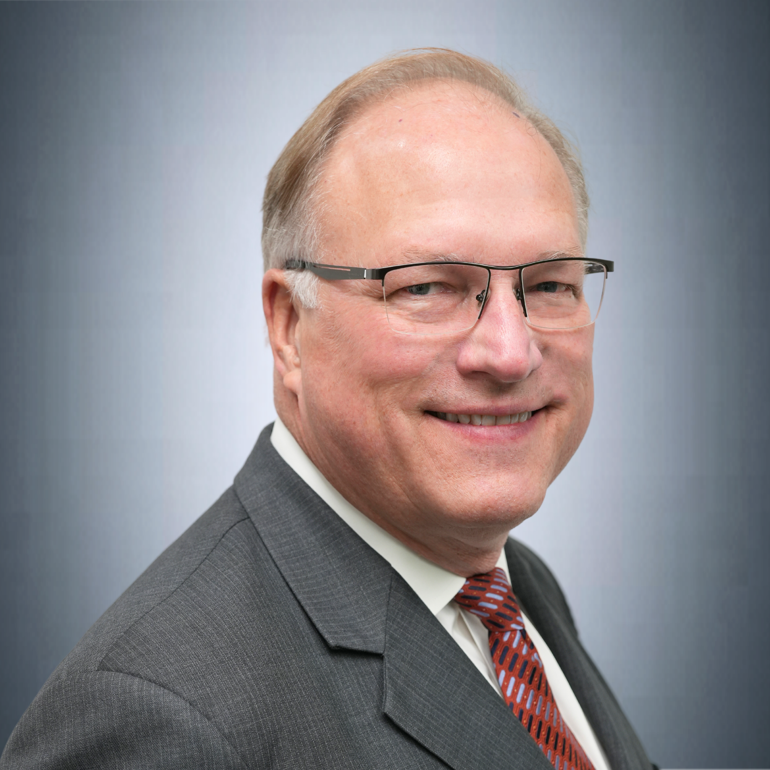 A middle-aged man with light skin, glasses, and short blond hair, wearing a dark suit, white shirt, and patterned red tie, smiling at the camera against a plain light blue background.