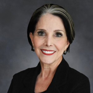 A professional woman with short dark hair streaked with gray, smiling, wearing a black blazer, and standing against a dark gray background.