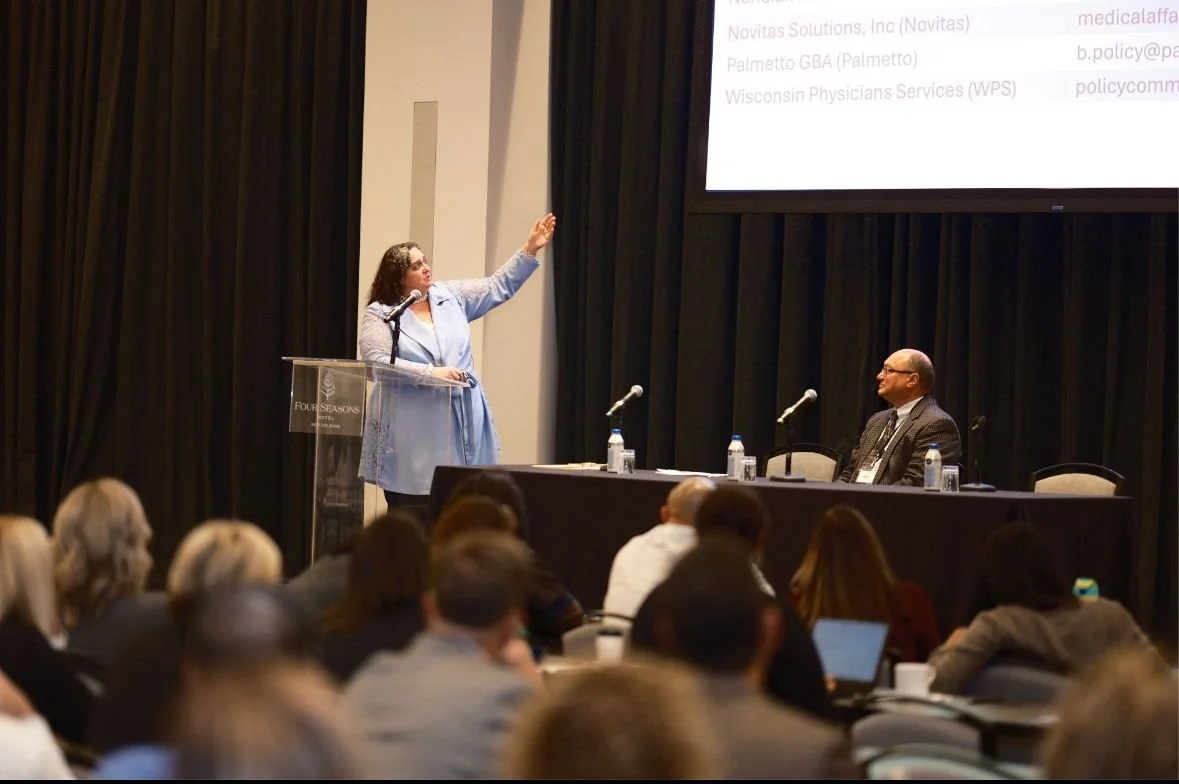 Woman giving a presentation on stage with a raised hand, seated man at a table with microphones and water bottles, audience listening at a conference.