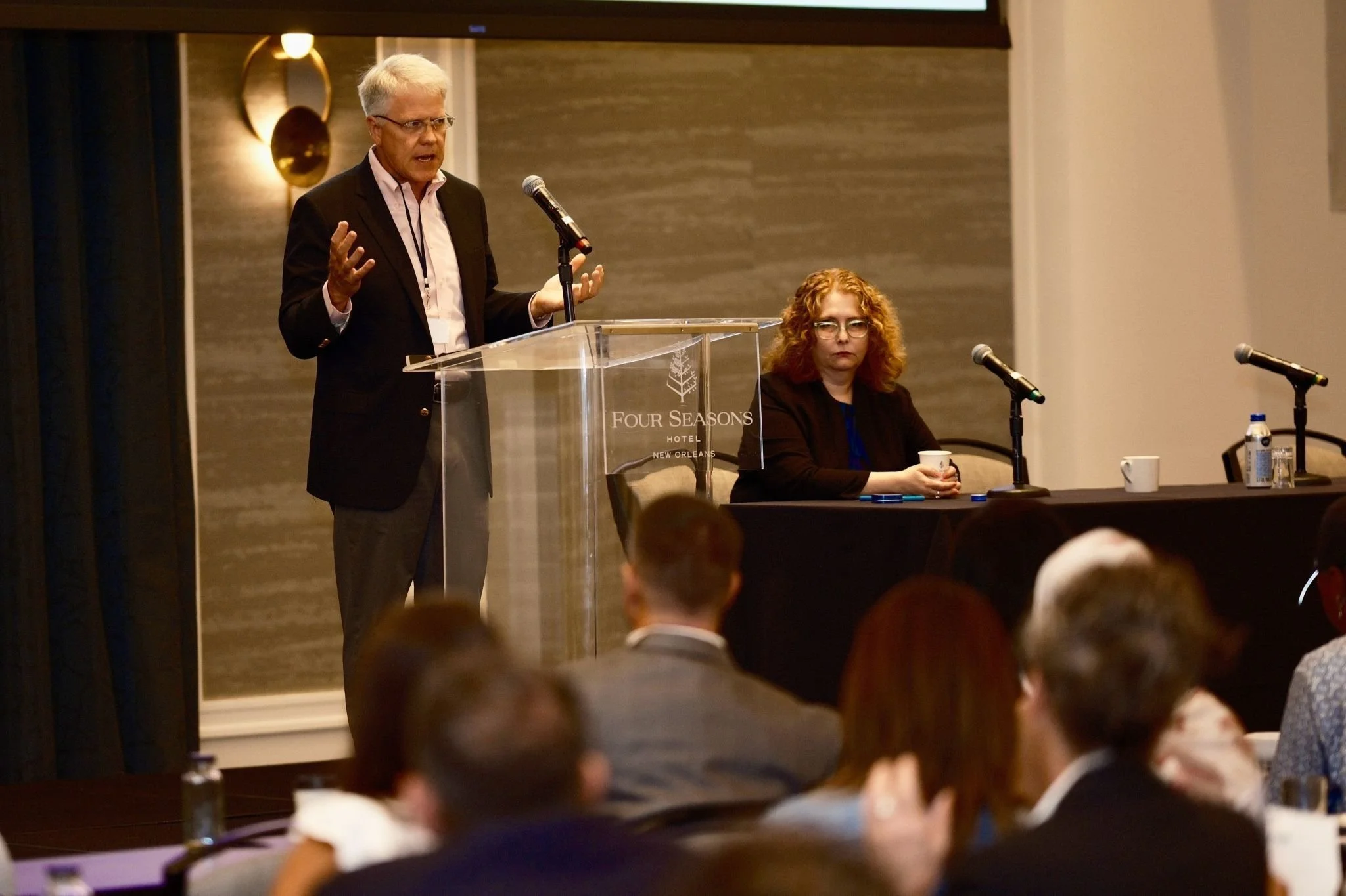 An older man with gray hair and glasses is speaking at a podium with a microphone at a conference in a hotel conference room, with a woman sitting at a table beside him and several attendees seated in front.