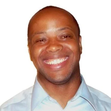A smiling man with a shaved head wearing a light blue dress shirt.