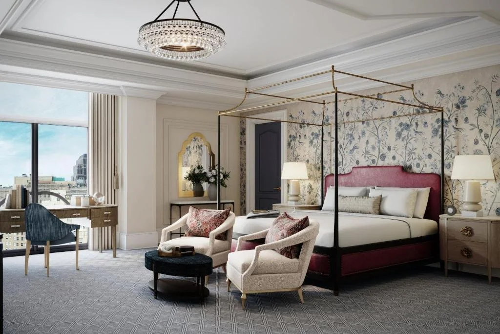 Elegant bedroom with four-poster bed, floral wallpaper, beige armchairs, a desk near the window, and a chandelier.
