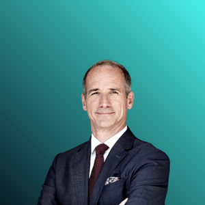 Professional middle-aged man in a dark suit with a white shirt and dark tie, standing with arms crossed in front of a gradient teal background. Dr Thomas Serena, Serena Group 
