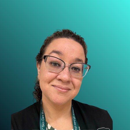 Close-up of a woman with curly hair, glasses, and a lanyard, smiling in front of a plain wall.