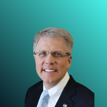 Professional headshot of a middle-aged man with gray hair, wearing glasses, a navy suit, white shirt, and light blue tie, against a blurred indoor background.