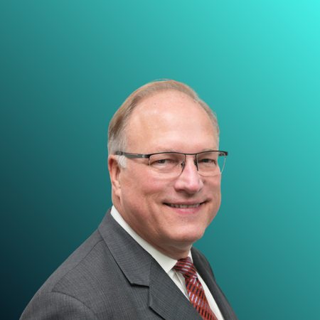 A middle-aged man with light skin, glasses, and short blond hair, wearing a dark suit, white shirt, and patterned red tie, smiling at the camera against a plain light blue background.