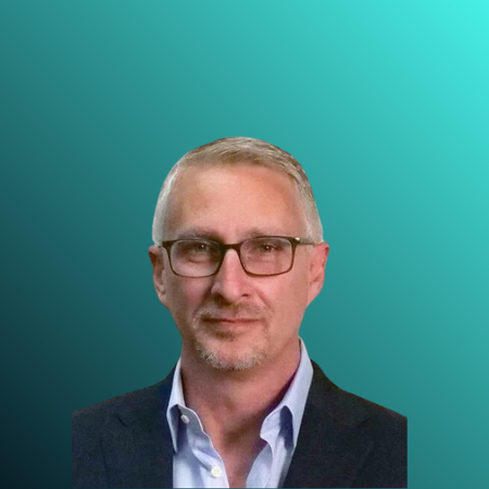 A middle-aged man with glasses, short gray hair, and light facial hair, wearing a dark blazer and a light blue collared shirt, standing indoors with a blurred office background.