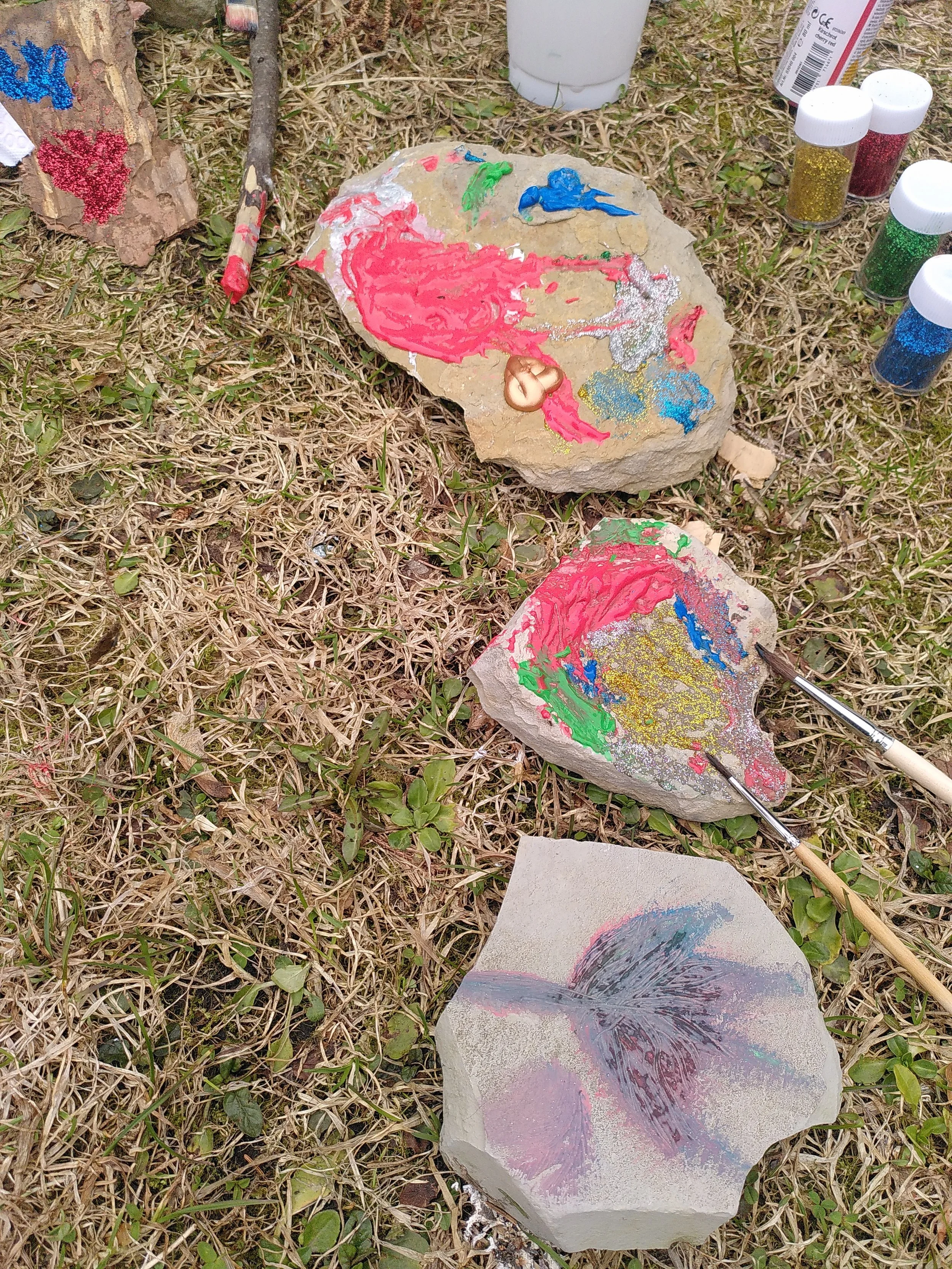 Steine, die bemalt wurden, einige mit bunten Farben und Glitzern, auf einem grassigen Boden. Es sind Malwerkzeuge und Glitzerfläschchen sichtbar.