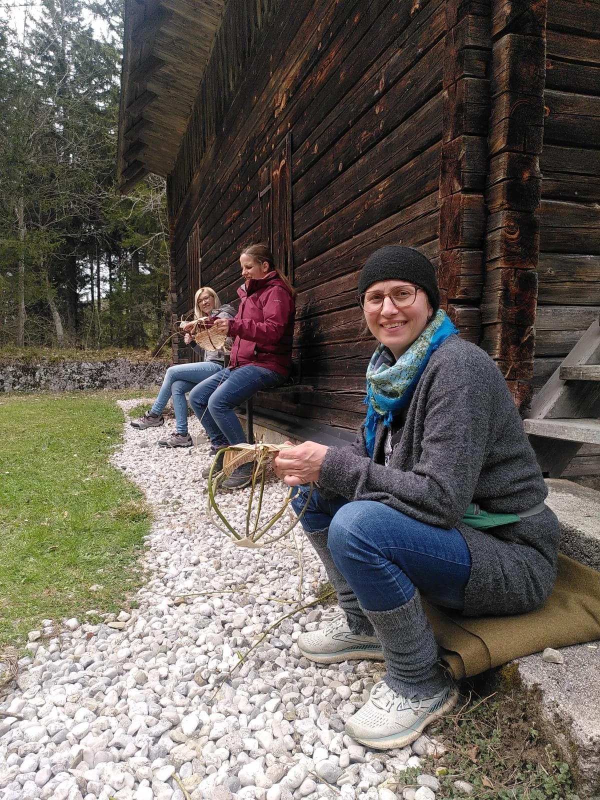 Drei Frauen sitzen an einem alten Holzgebäude im Freien, die vordere trägt eine Mütze und eine Regenjacke, die mittlere ist beim Basteln und die hintere liest. Alle scheinen beim Handwerk oder beim Entspannen im Grünen.