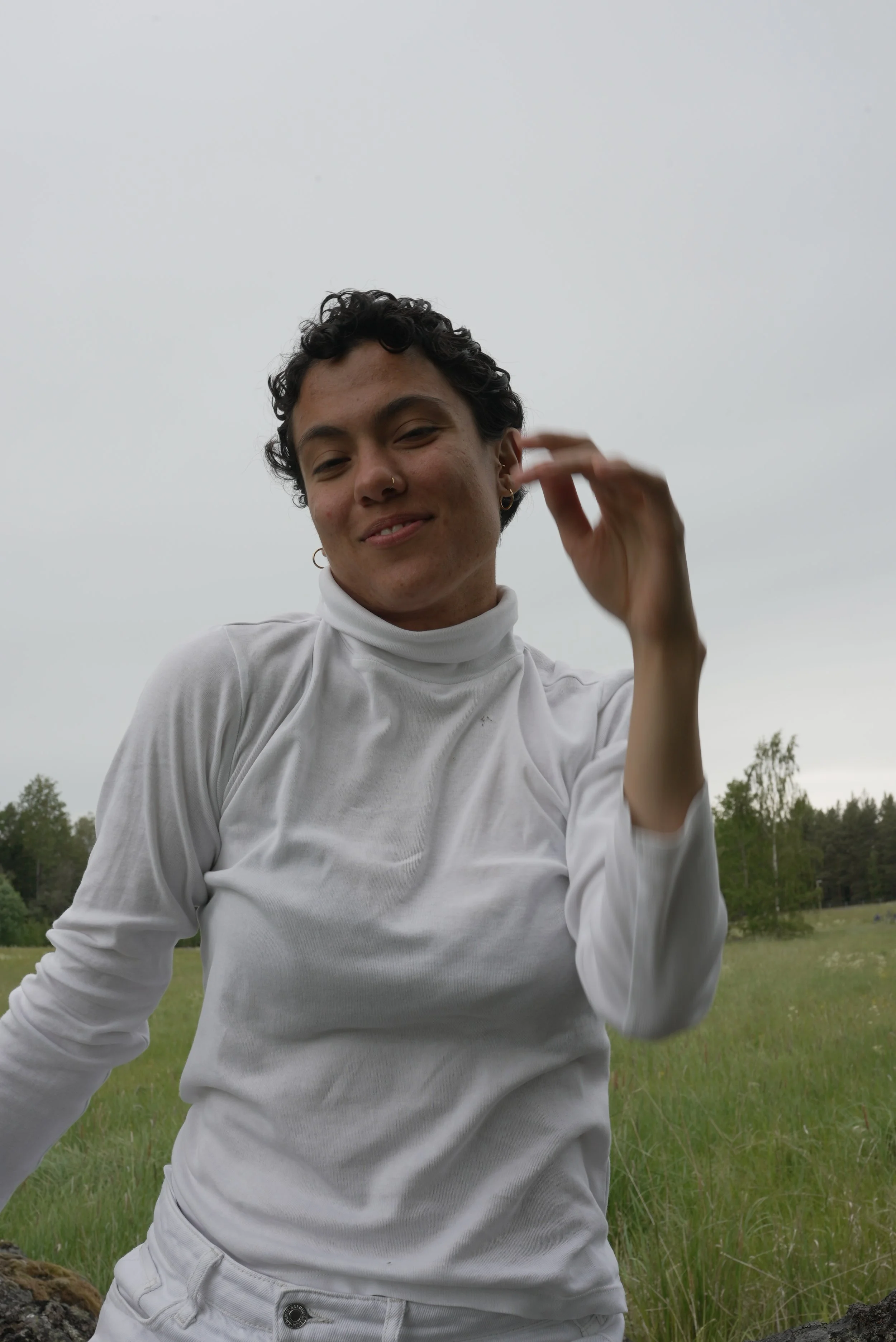 A person with short dark curly hair, wearing a white turtleneck and pants, smiling outdoors on a cloudy day with green grass and trees in the background. House dancer, Hip Hop dancer, contemporary dancer. Practicing awareness and community.