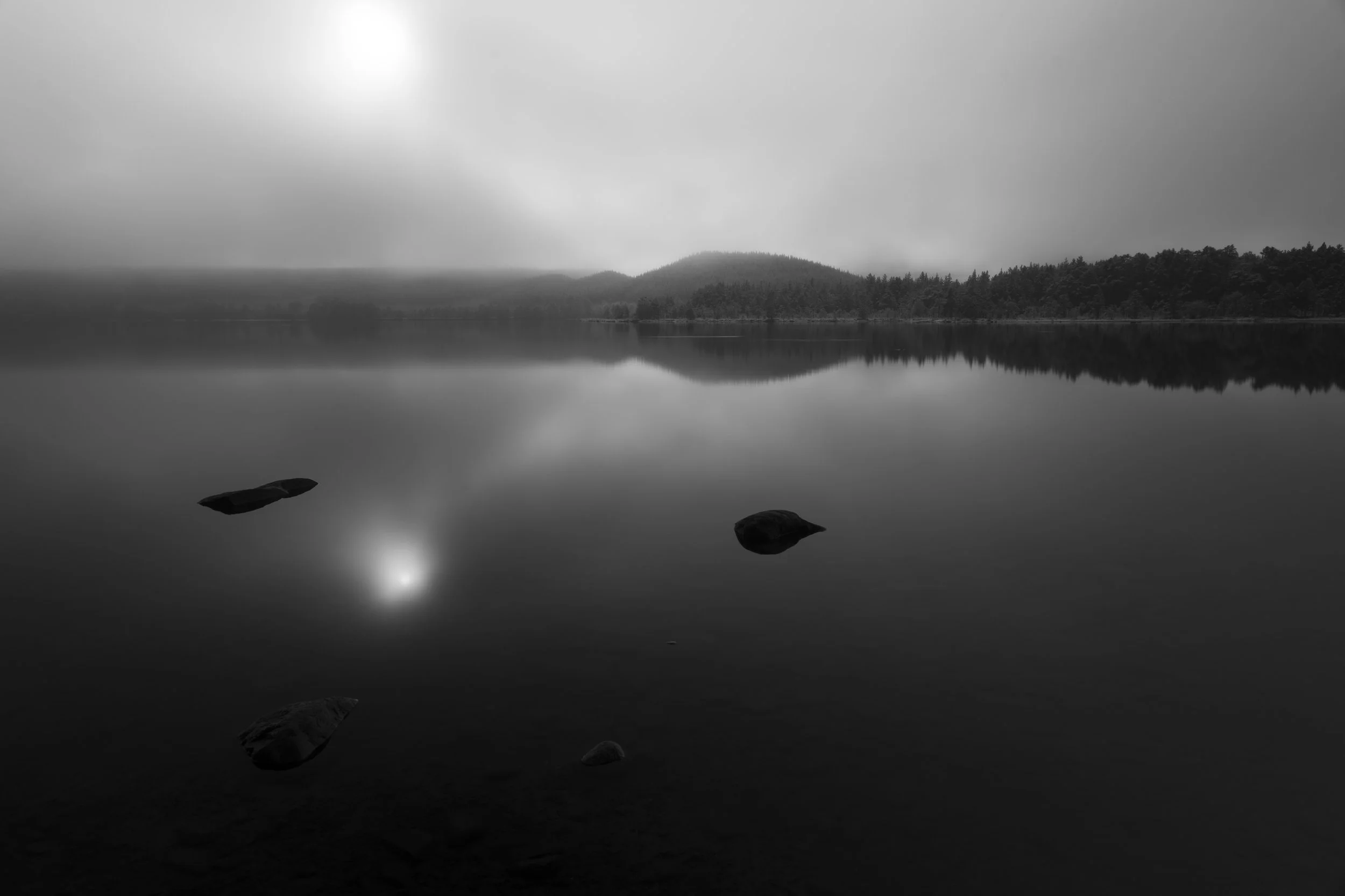 Black and white photo of a calm lake with four rocks near the shore, surrounded by forested hills under an overcast sky.
