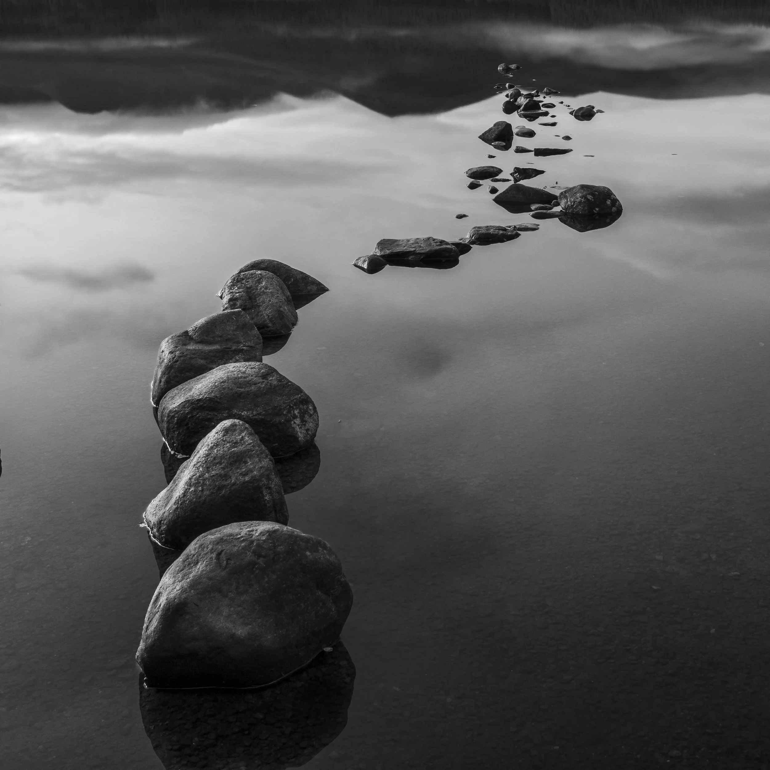 A black and white photo of a line of rocks partially submerged in calm water, creating a pathway leading into the distance with mountains and a cloudy sky reflected in the water.