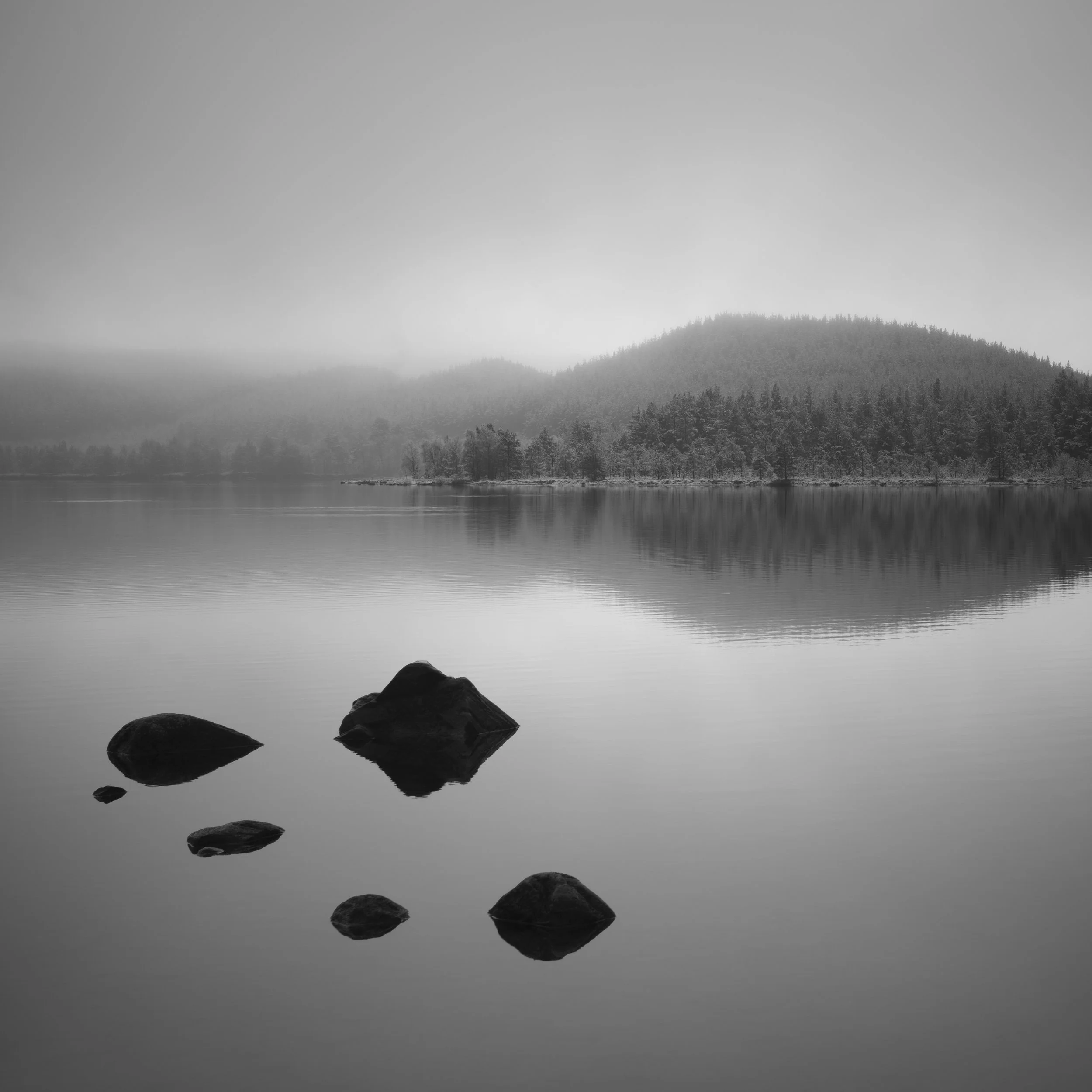 Black and white photograph of a calm lake with rocks at the foreground, reflecting trees and mountainous terrain in the background, under a hazy sky.