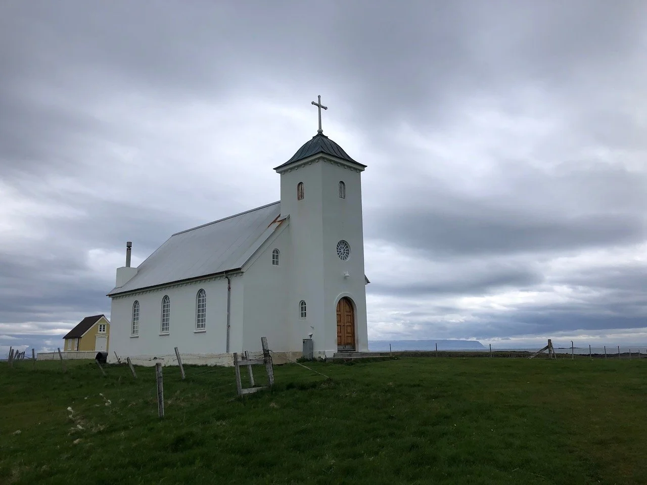 A photo of a simple white church. Green grass is in the foreground. A foreboding gray sky is overhead. Photo credit: Kelly Nielsen